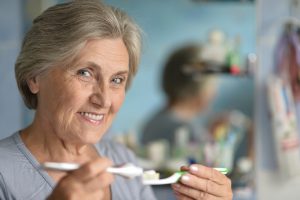  An older woman is brushing her teeth with a toothbrush, demonstrating good oral hygiene in a bright bathroom setting.