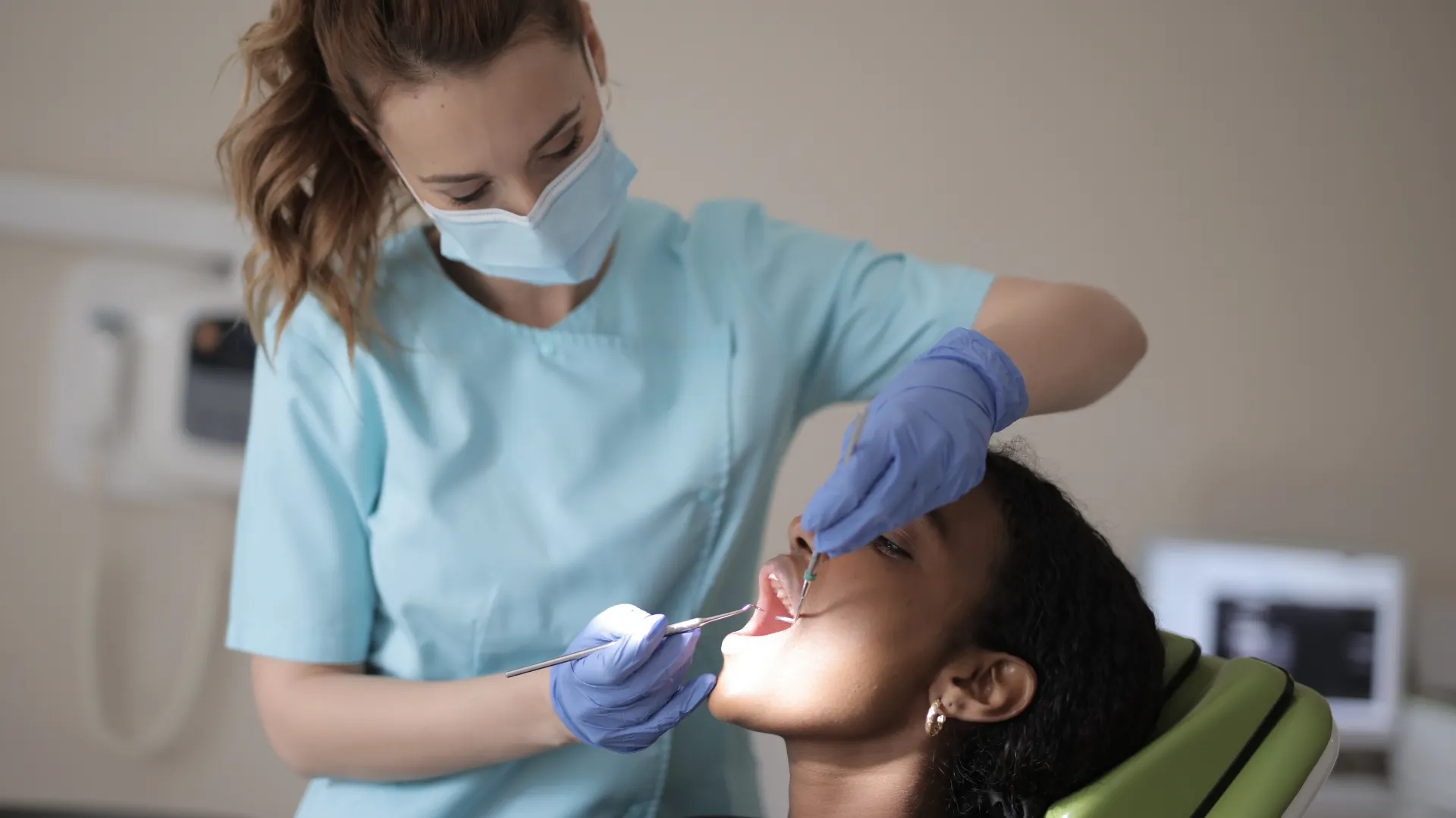 A female dentist or dental hygienist wearing a blue uniform, mask, and gloves examines a patient's teeth using a dental mirror and probe in a clinical setting.