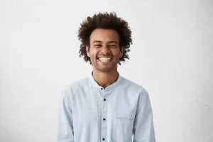 a smiling man with curly hair wearing a light-colored button-up shirt against a plain white background. His bright and cheerful expression conveys happiness and positivity.