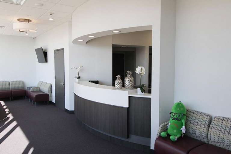 A modern dental office reception area featuring a curved front desk, seating with cushioned benches, and a green plush toy on one of the chairs