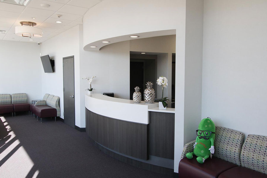 A modern dental office reception area featuring a curved front desk, seating with cushioned benches, and a green plush toy on one of the chairs