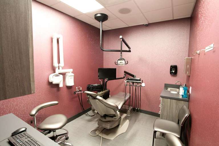 A dental exam room with pinkish-red walls, featuring a dental chair, overhead examination light, mounted X-ray equipment, and a workstation with a monitor and countertop.