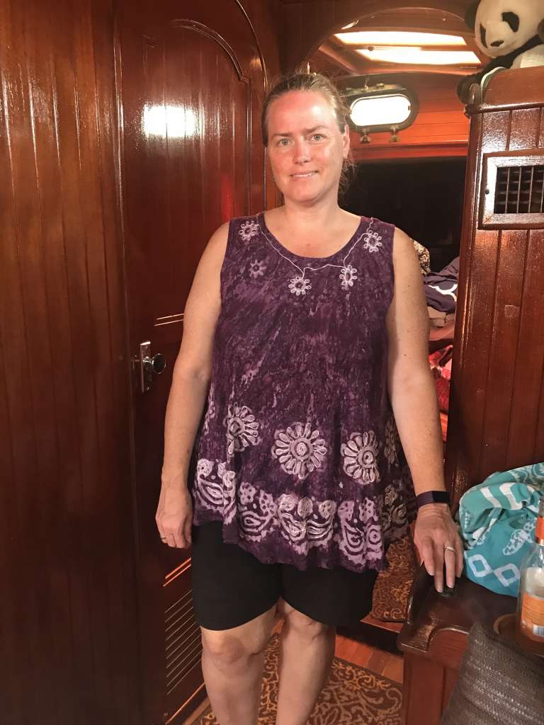 A woman in a purple floral top stands inside a wooden-paneled room, smiling at the camera.