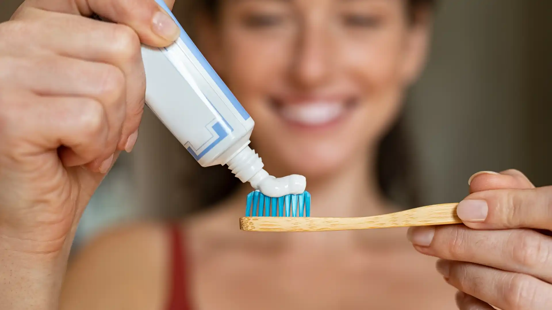 Close-up of toothpaste being applied to a toothbrush with a smiling woman in the background."
