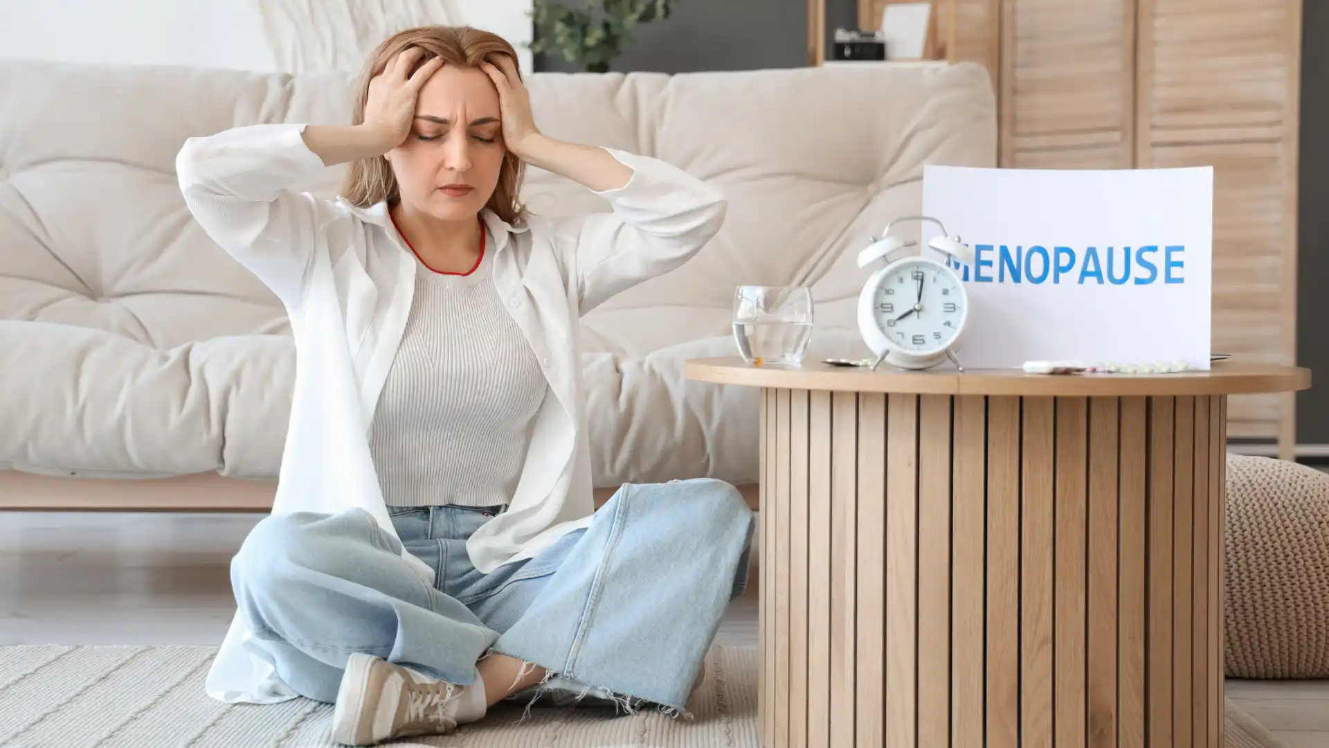 Stressed woman sitting on the floor, holding her head, with an alarm clock and papers beside her.