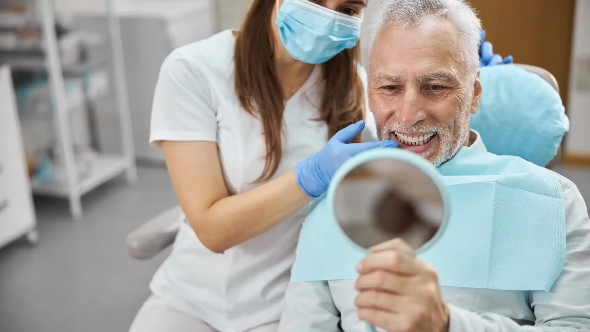 Elderly man at a dental clinic smiling while looking at his reflection in a handheld mirror, assisted by a masked dental professional