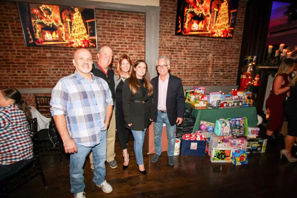 A group of five people posing in a dimly lit venue with exposed brick walls, with a table filled with colorful gift donations in the background.