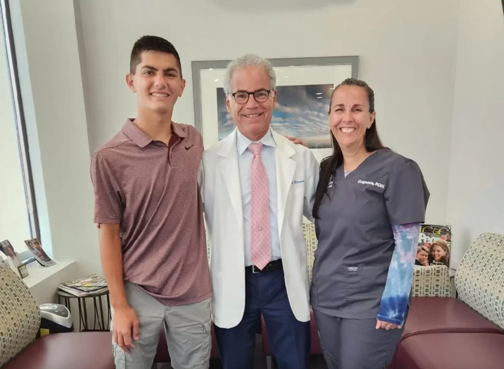 Three smiling people posing in a professional office, including a doctor in a white coat and two others in casual and medical attire.