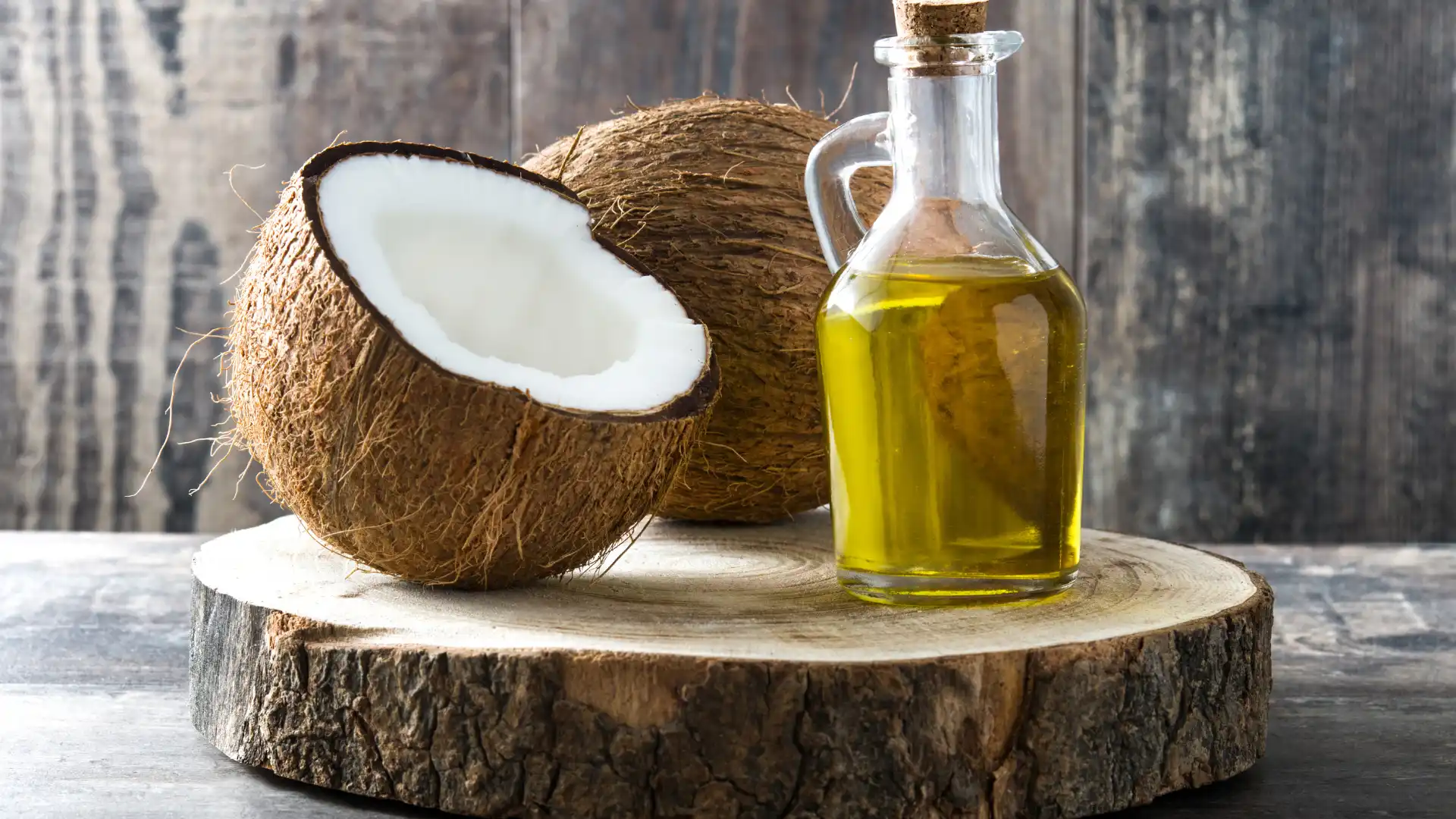 Glass bottle of coconut oil with fresh coconuts on a wooden surface.