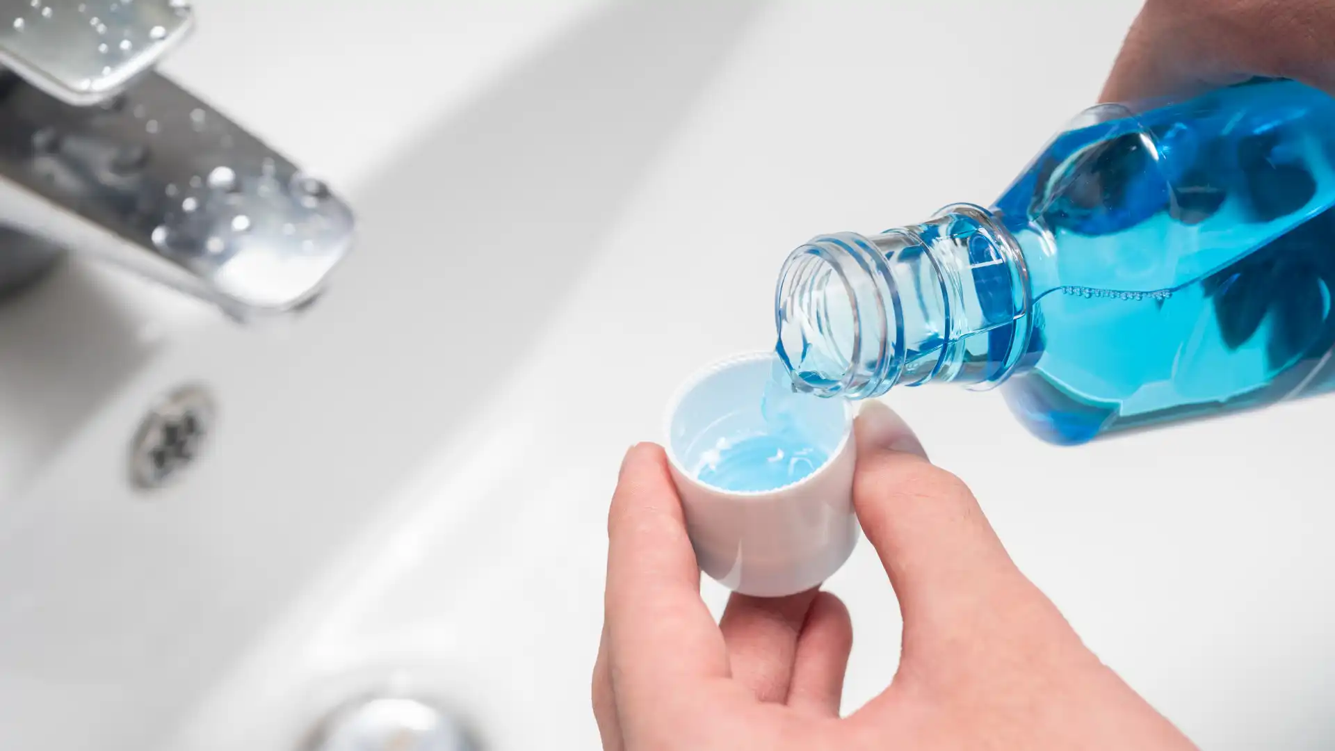 Person pouring blue mouthwash into a cap over a bathroom sink.