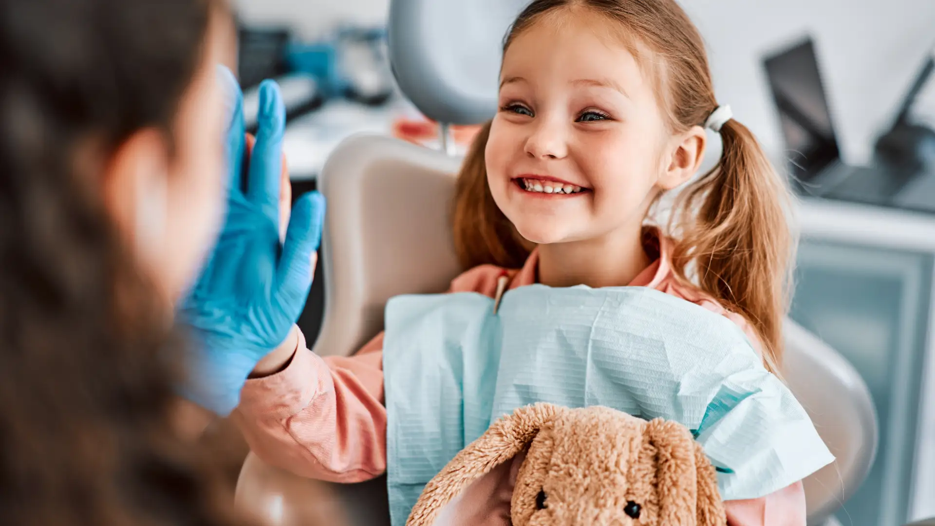 Happy young girl in a dental chair giving a high-five to a gloved dentist while holding a stuffed bunny.