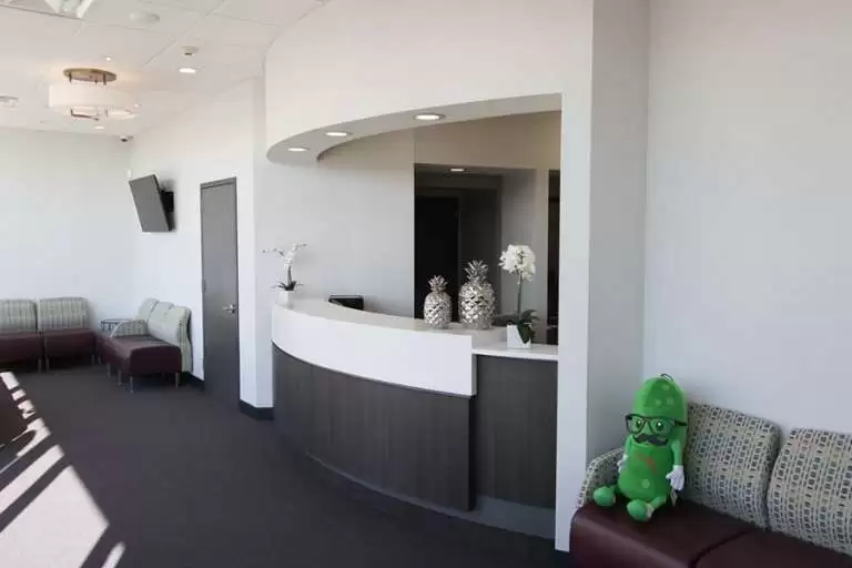 A modern dental office reception area featuring a curved front desk, seating with cushioned benches, and a green plush toy on one of the chairs