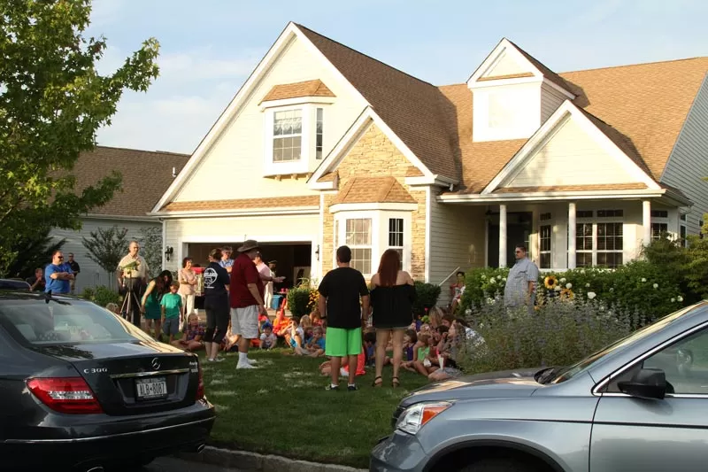 A gathering of people outside a suburban house during daytime, with cars parked in the foreground.