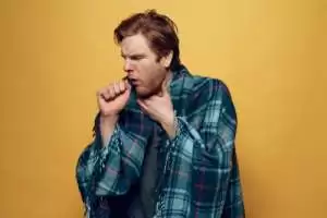Sick man wrapped in a plaid blanket, coughing into his hand against a yellow background.