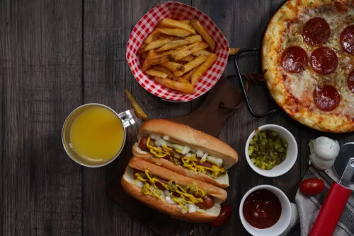 Top-down view of a meal featuring two hot dogs with mustard and onions, a basket of French fries, a pepperoni pizza, a glass of orange juice, and condiments on a rustic wooden table.