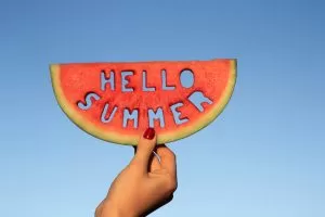 A hand holds a slice of watermelon featuring the phrase "Hello Summer" written on it.