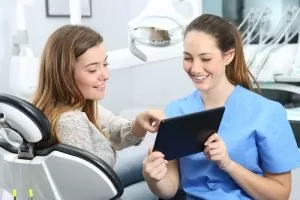 A dental professional in blue scrubs showing a tablet to a smiling patient in a modern clinic.