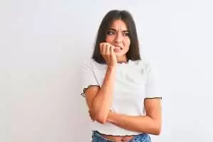 A young woman in a white top looks anxious, biting her nails with crossed arms.