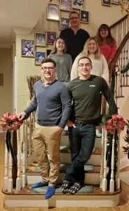 A group of six people posing on a decorated staircase in a warmly lit home, with framed photos on the wall behind them.