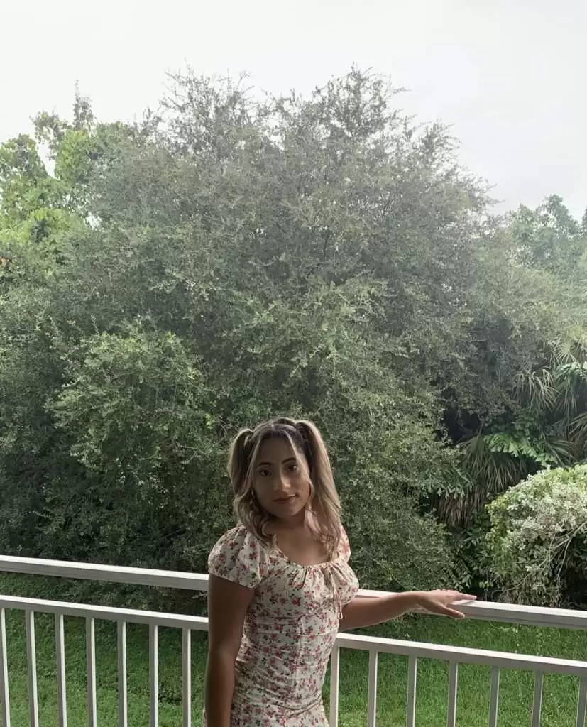 a young woman with pigtails wearing a floral dress, standing by a white railing with lush green trees in the background