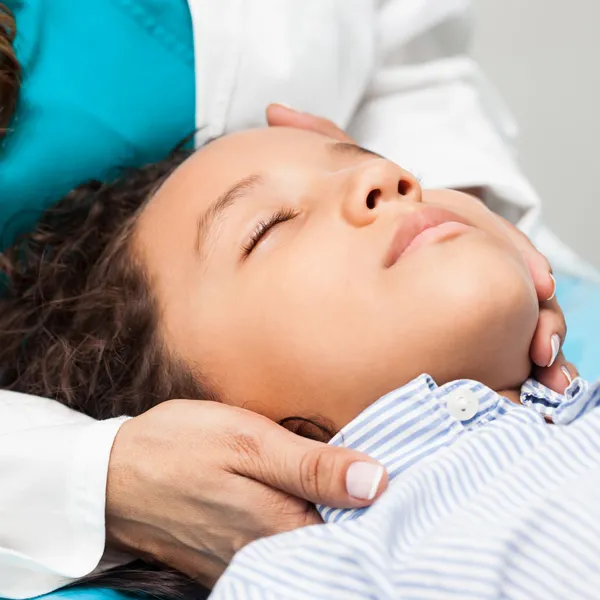 A child lies back in a dental chair with eyes closed while a dentist supports the child’s head during an exam.